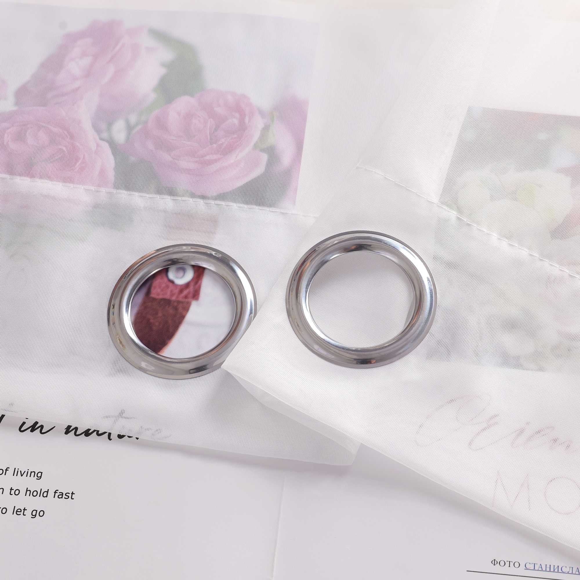 Der weiße Vorhangstoff auf dem Bild ist mit zwei Metalllöchern versehen. Durch den hellen und transparenten Stoff ist ein florales Muster im Hintergrund schwach zu erkennen. So wie es aussieht, sind die Ösen wahrscheinlich aus rostfreiem Stahl gefertigt.