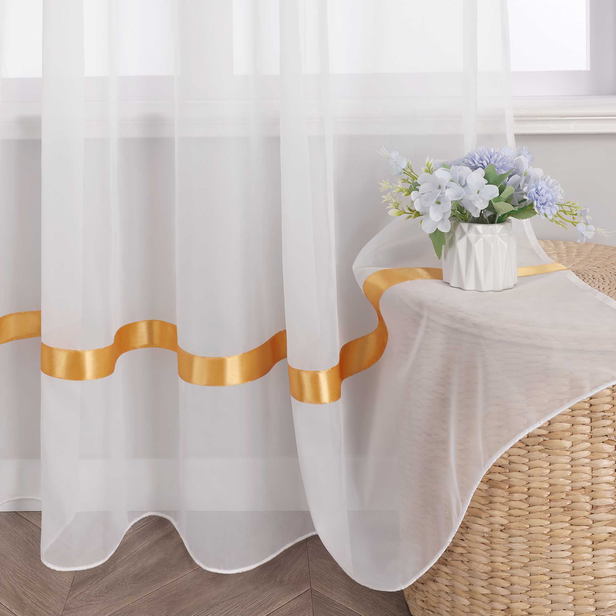 Am Fenster haengt ein weisser, halbtransparenter Vorhang mit orangefarbenen Satinbaendern aus Voile.  Darauf steht eine weisse Vase mit einem Strauss aus blau-weissen Blumen.