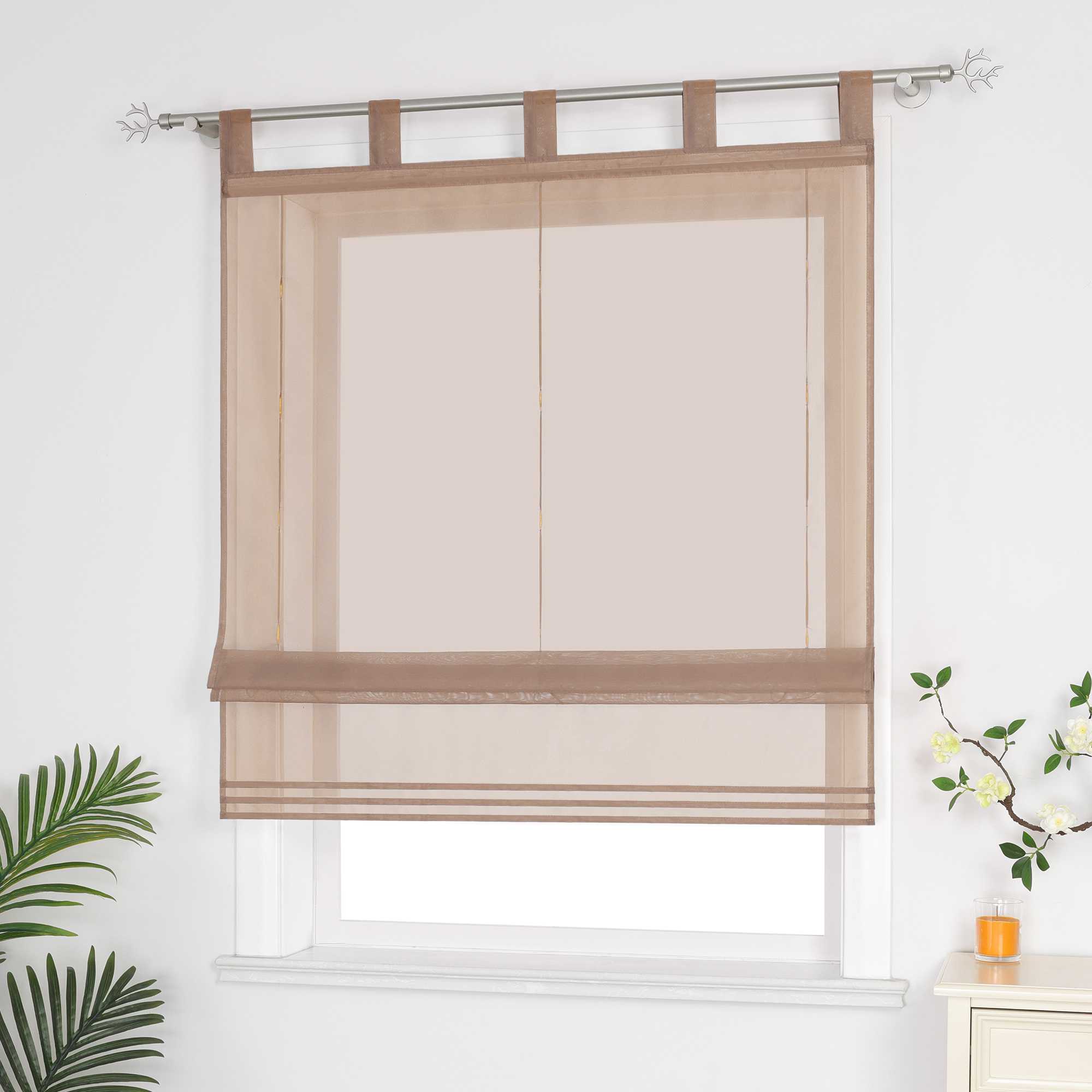 Halbtransparenter brauner Raffrollo hängt vor dem Fenster und sorgt für eine helle, luftige Atmosphäre. Daneben setzen grüne Pflanzen dekorative Akzente. Seitliche Ansicht.
