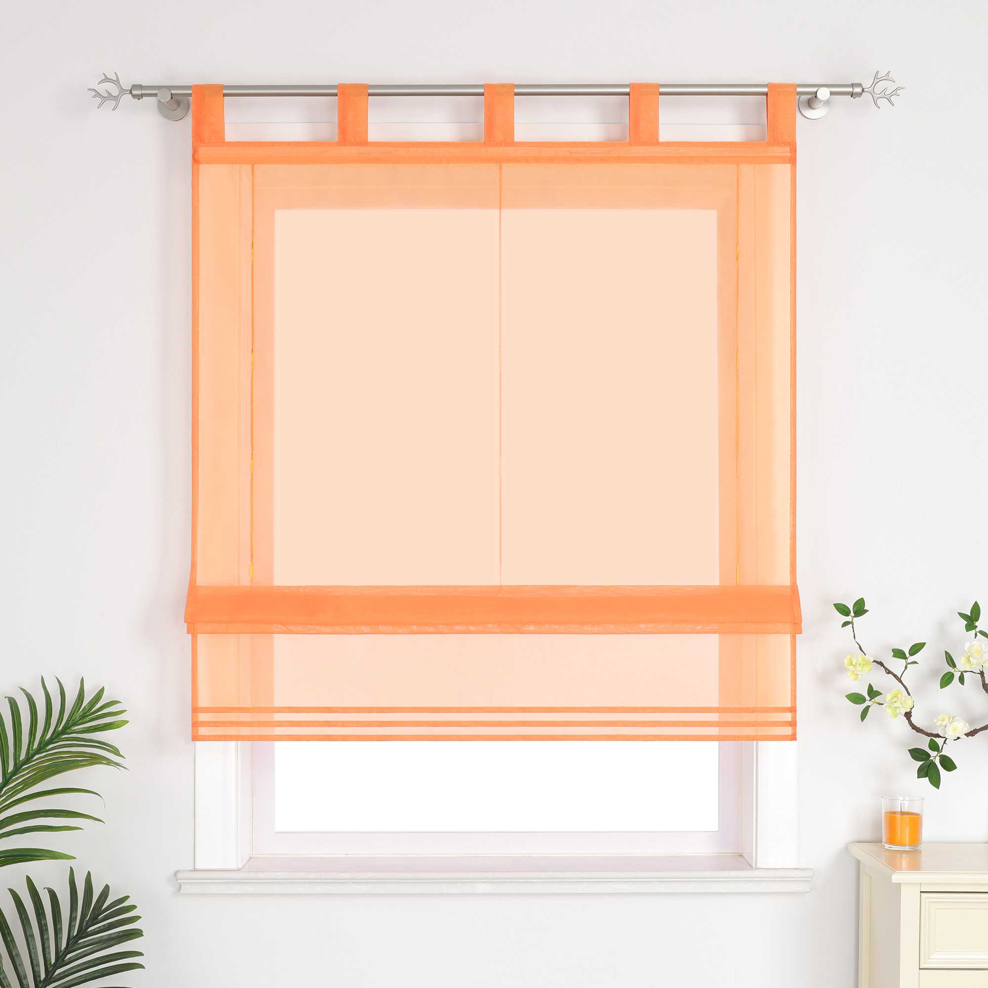 Im Wohnzimmer hängen orangefarbene Rollos. Daneben sind dekorative Grünpflanzen und Duftkerzen aufgestellt.