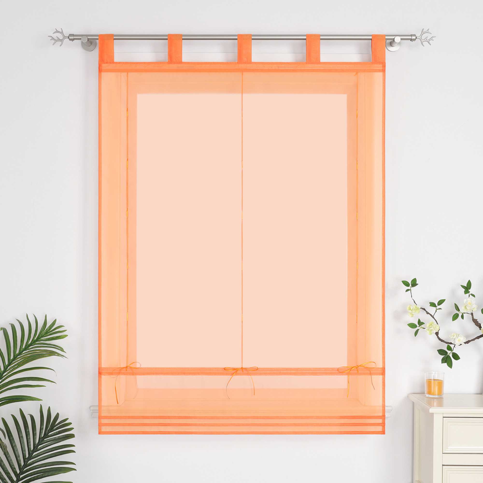 Im Wohnzimmer hängen orangefarbene Rollos. Neben ihnen stehen dekorative Grünpflanzen und Duftkerzen.