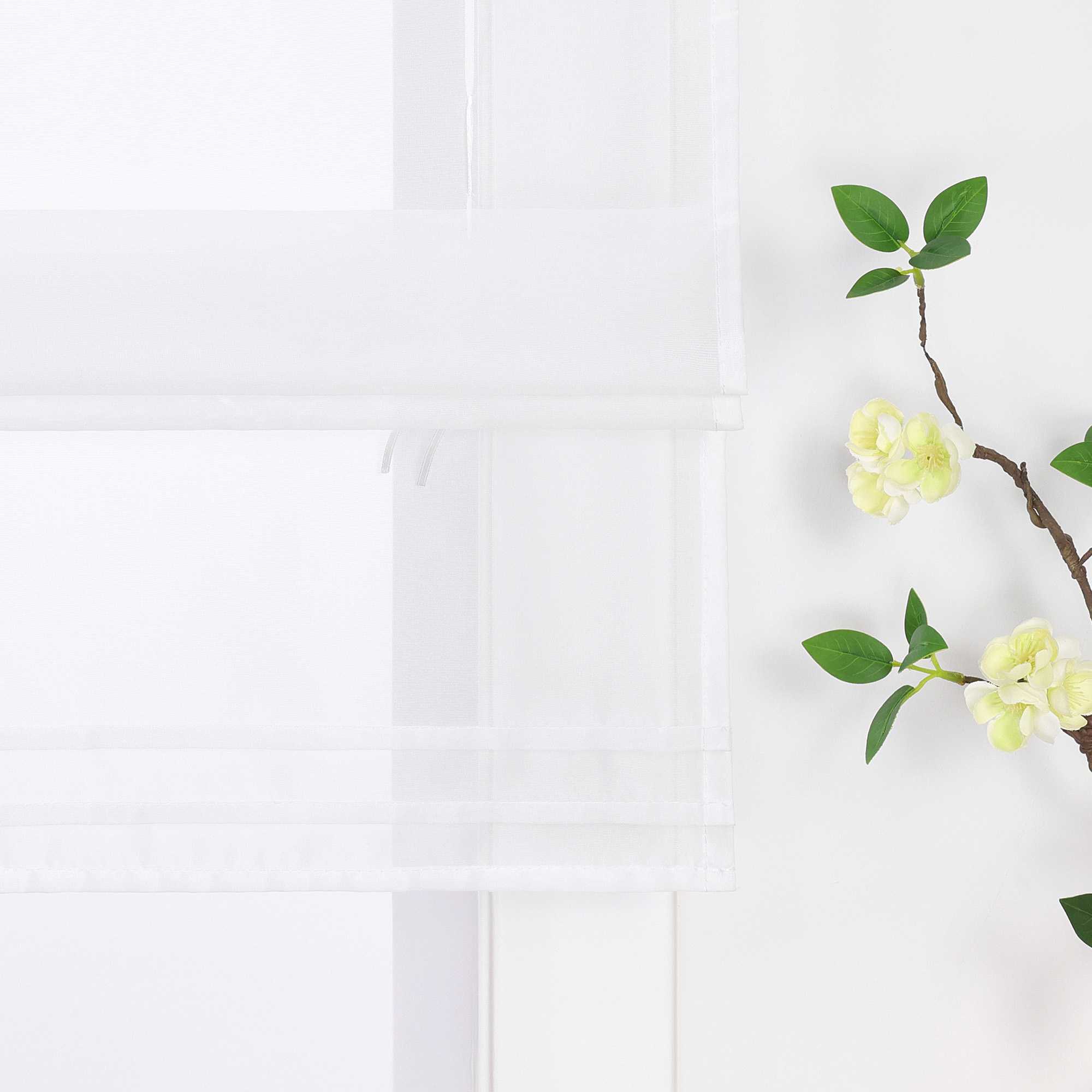 Detailansicht des unteren Bereichs eines halbtransparenten weißen Raffrollos. Der Stoff ist sanft hochgezogen und bildet dekorative Falten. Eine stilvolle Blumen-Deko neben dem Fenster rundet das Gesamtbild harmonisch ab.