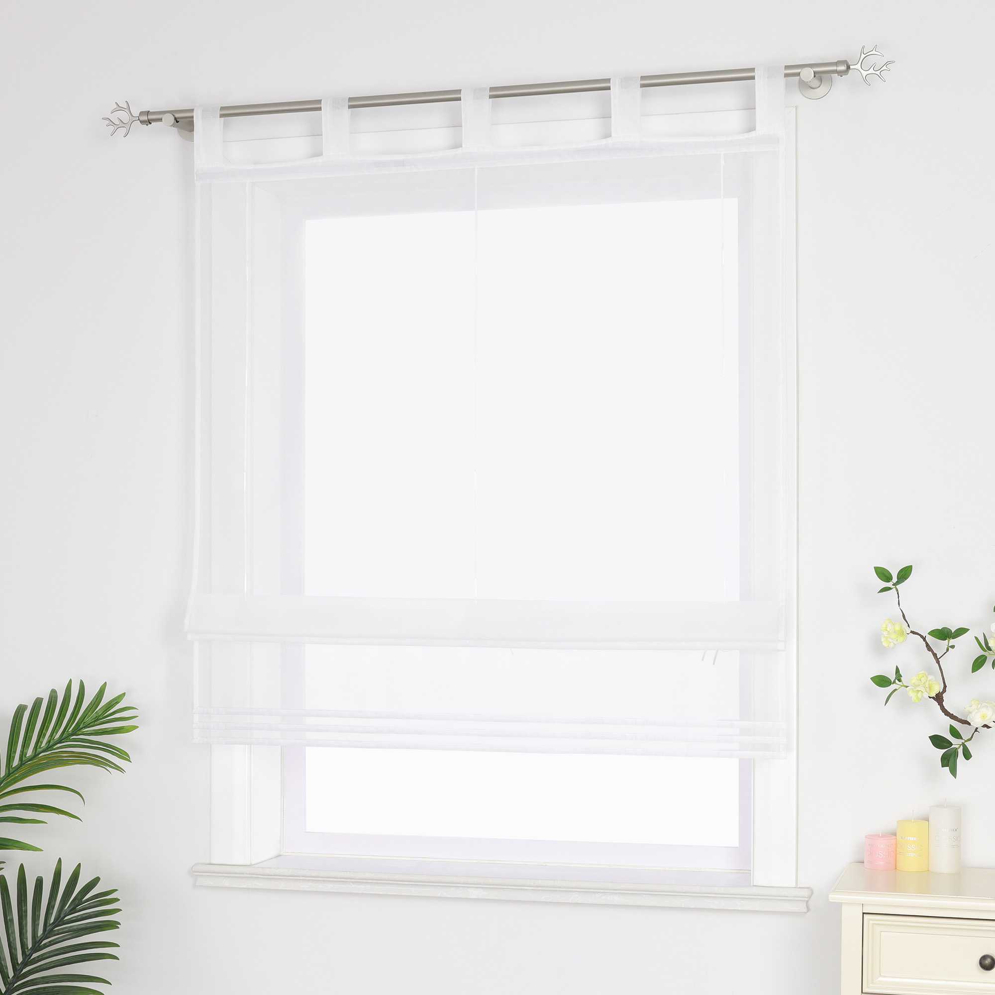 Halbtransparenter weißer Raffrollo hängt vor dem Fenster und sorgt für eine helle, luftige Atmosphäre. Daneben setzen grüne Pflanzen dekorative Akzente. Seitliche Ansicht.