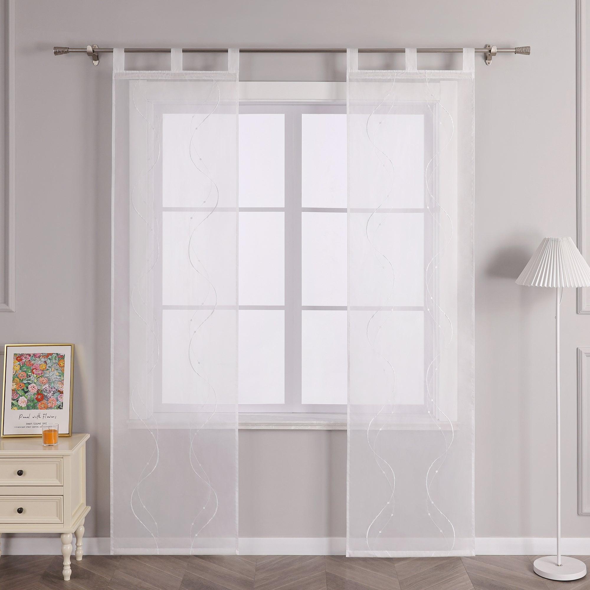 Vor einer hellgrauen Wand befindet sich ein Fenster, an dem eine weisse transparente Schiebegardine mit Wellenperlenmuster und Stickerei haengt. Die Gardine ist mit Schlaufen an einer Metallstange befestigt. Links vom Fenster steht ein heller Schrank mit Schubladen, auf dem ein buntes Blumenbild und ein kleines oranges Objekt platziert sind. Rechts vom Fenster steht eine Stehlampe mit einem weissen gestreiften Lampenschirm.