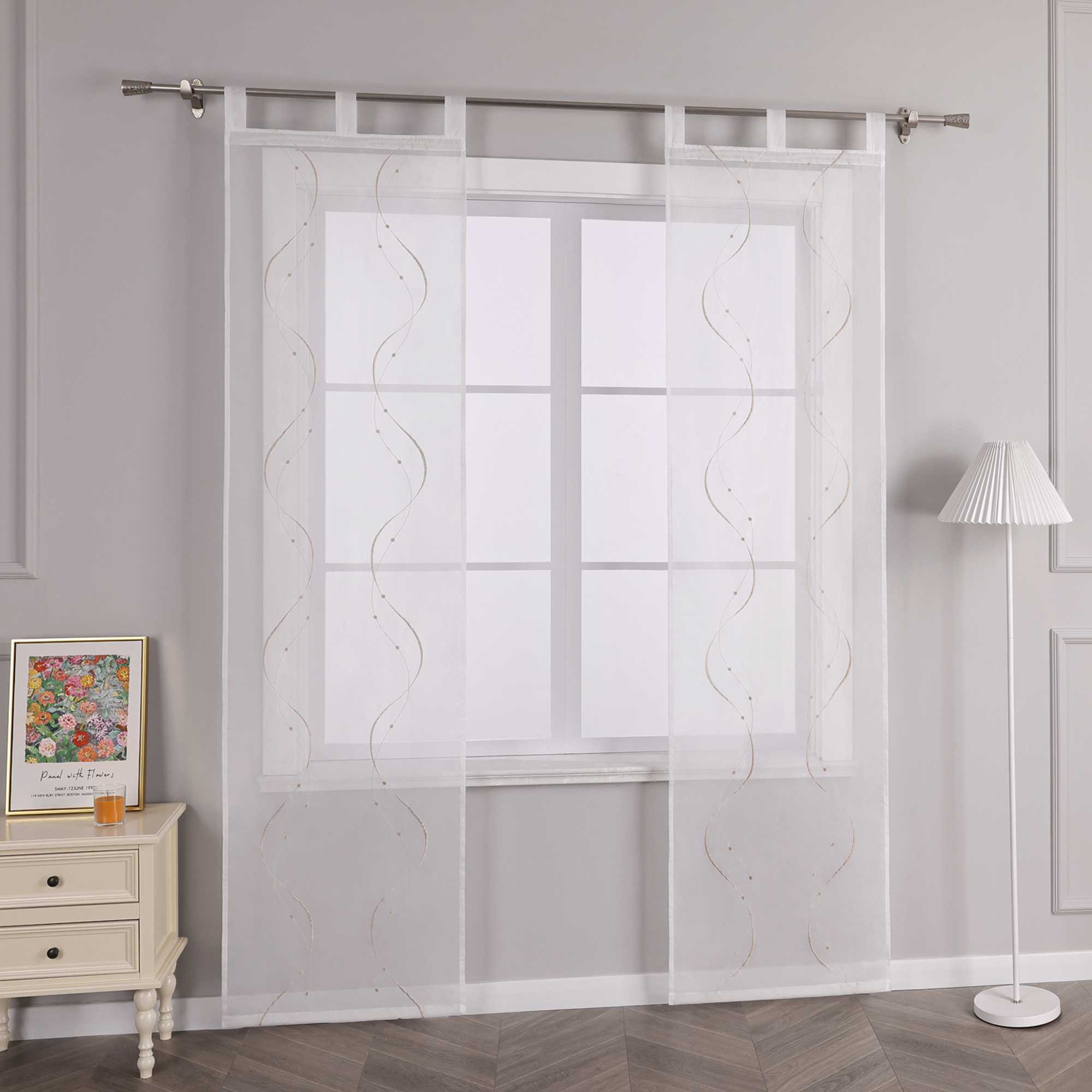 Joyswahl Das Bild zeigt eine Seitenansicht der weissen transparenten Gardine, die an einer Metallstange hängt, mit sandfarbenen Wellenperlenmustern und Stickerei. Links vom Fenster steht ein heller Schrank mit Schubladen, auf dem ein farbenfrohes Blumenbild und ein orangenes kleines Objekt stehen. Rechts vom Fenster befindet sich eine Stehlampe mit einem weissen Streifenlampenschirm.