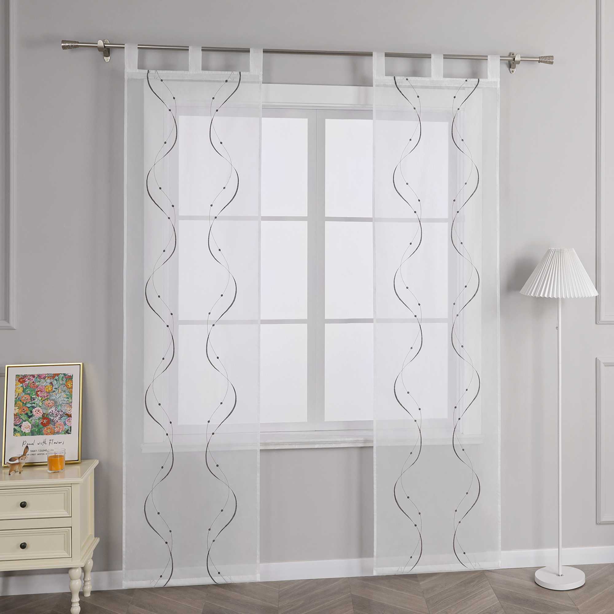 Joyswahl – Das Bild zeigt die Seitenansicht einer weissen transparenten Gardine mit Wellenperlenmuster und Stickerei, die an einer Metallstange haengt. Links vom Fenster steht ein heller Schubladenschrank mit einem farbigen Blumenbild und einem kleinen orangen Objekt darauf. Rechts vom Fenster steht eine Stehlampe mit einem weissen gestreiften Lampenschirm.