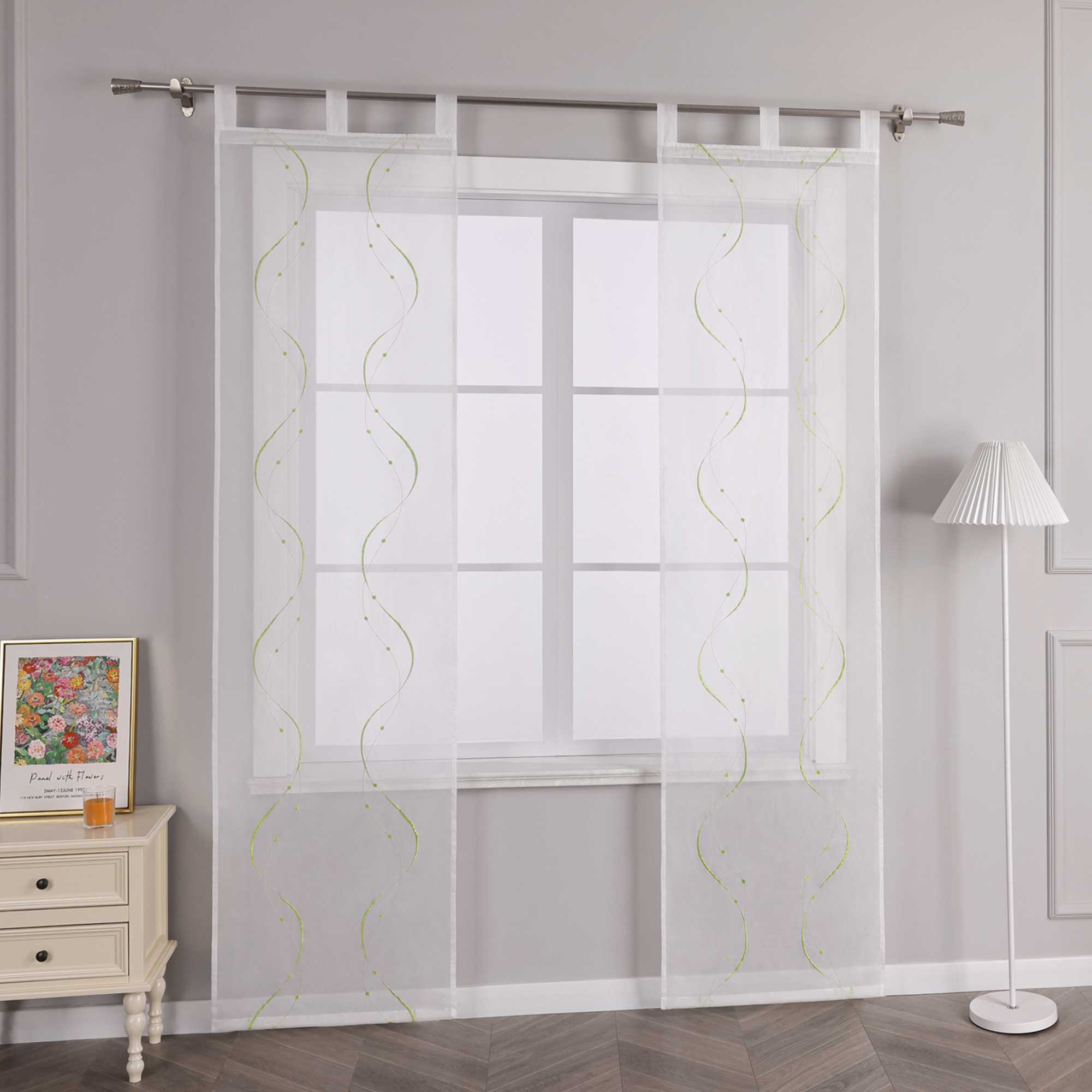Joyswahl – Das Bild zeigt die Seitenansicht einer weissen transparenten Gardine mit grünem Wellenperlenmuster und Stickerei, die an einer Metallstange haengt. Links vom Fenster steht ein heller Schubladenschrank mit einem farbigen Blumenbild und einem kleinen orangen Objekt darauf. Rechts vom Fenster steht eine Stehlampe mit einem weissen gestreiften Lampenschirm.