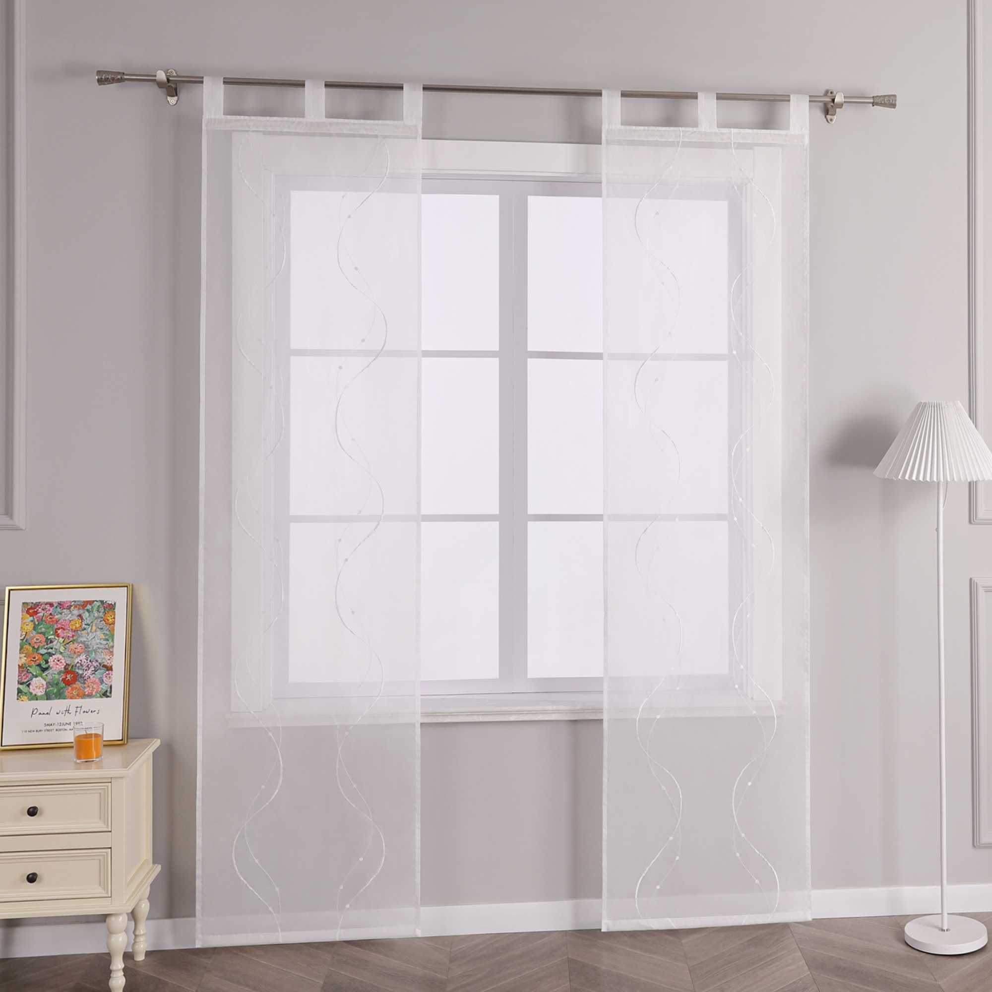 Das Bild zeigt die Seitenansicht einer weissen transparenten Gardine mit Wellenperlenmuster und Stickerei, die an einer Metallstange mit Schlaufen haengt. Links vom Fenster steht ein heller Schrank mit Schubladen, auf dem ein buntes Blumenbild und ein kleines oranges Objekt platziert sind. Rechts vom Fenster befindet sich eine Stehlampe mit einem weissen gestreiften Lampenschirm.