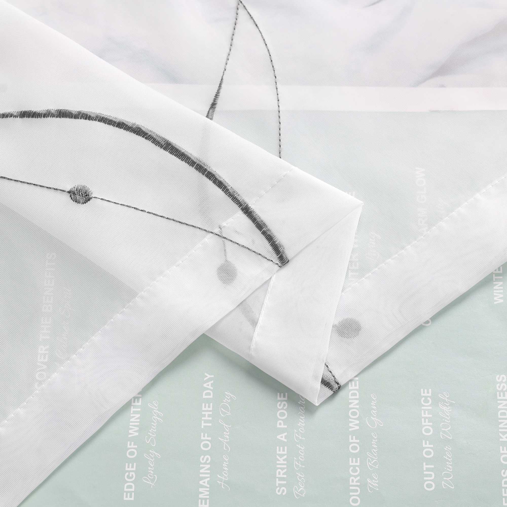 Ein weisser, duenn gewebter Stoff mit schmalem Wellenlinienmuster ist mehrfach gefaltet. Im Hintergrund befindet sich Papier mit aufgedrucktem Text und Motiven.