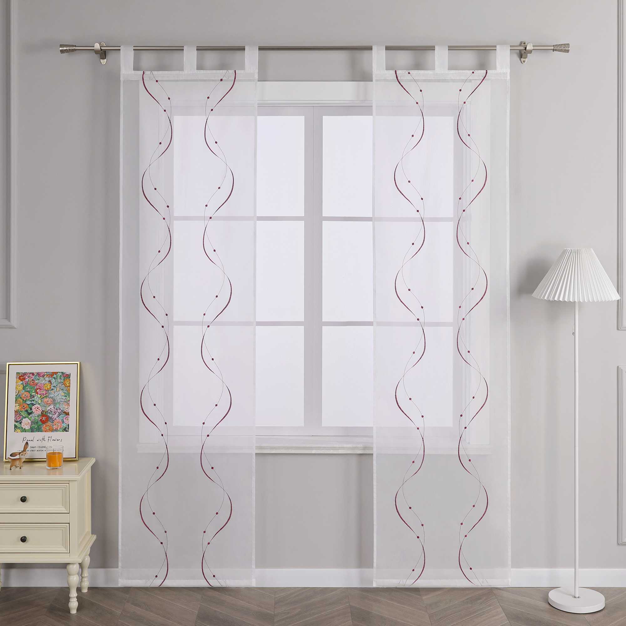 Vor einer hellgrauen Wand befindet sich ein Fenster, an dem eine weisse transparente Schiebegardine haengt. Die Gardine ist mit symmetrischen lila wellenfoermigen Perlenmustern und Stickereien versehen. Sie ist an einer Metallstange mit Schlaufen befestigt. Links vom Fenster steht ein heller Schrank mit Schubladen, auf dem ein farbenfrohes Blumenbild und ein orangefarbenes Objekt platziert sind. Rechts vom Fenster steht eine Stehlampe mit einem weiss gestreiften Lampenschirm.