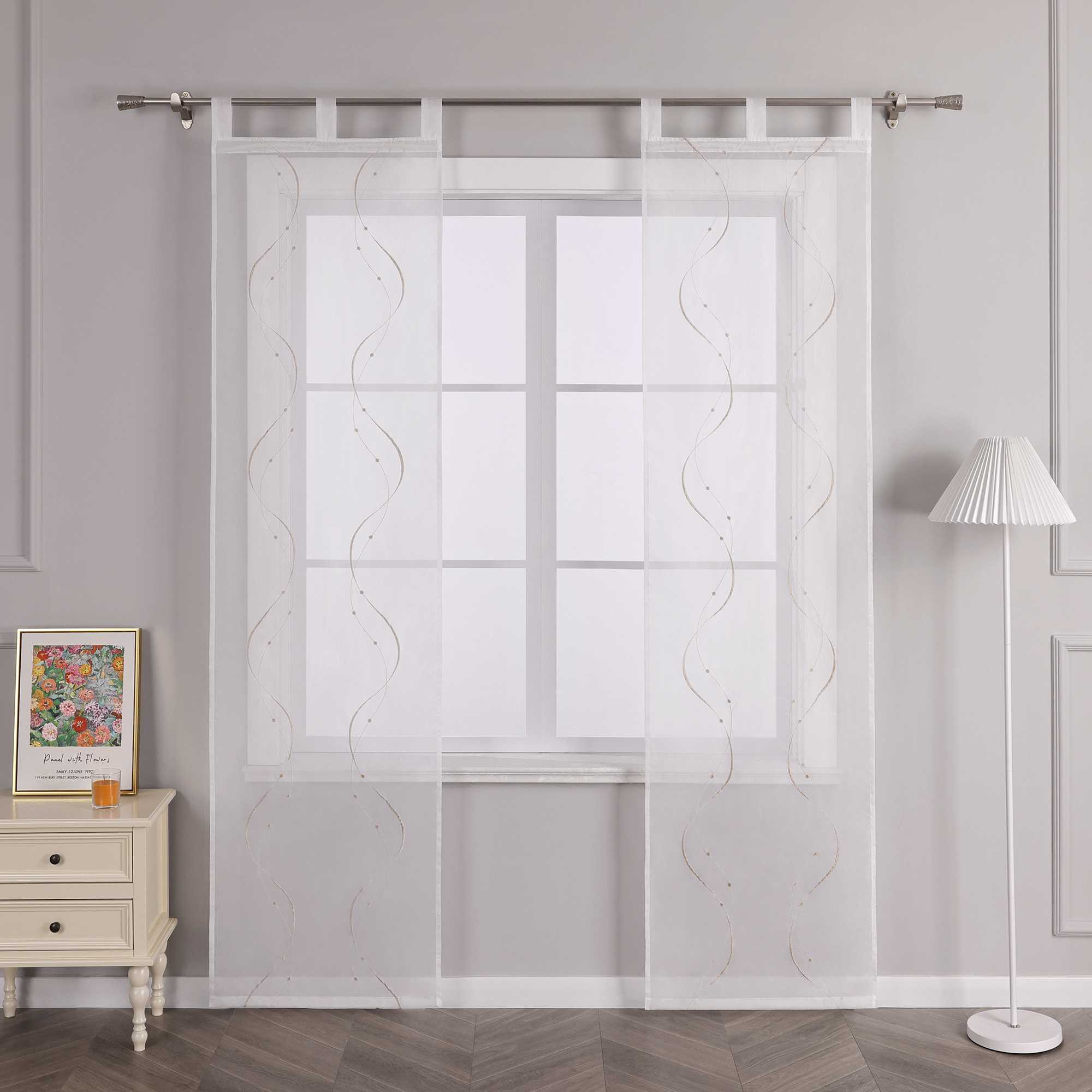 Vor einer hellgrauen Wand befindet sich ein Fenster, an dem eine weisse transparente Schiebe-Gardine mit sandfarbenen symmetrischen Wellenperlenmustern und Stickerei hängt. Die Gardine ist an einer Metallstange mit Schlaufen befestigt. Links vom Fenster steht ein heller Schrank mit Schubladen, auf dem ein farbenfrohes Blumenbild und ein orangenes kleines Objekt stehen. Rechts vom Fenster befindet sich eine Stehlampe mit einem weissen Streifenlampenschirm.
