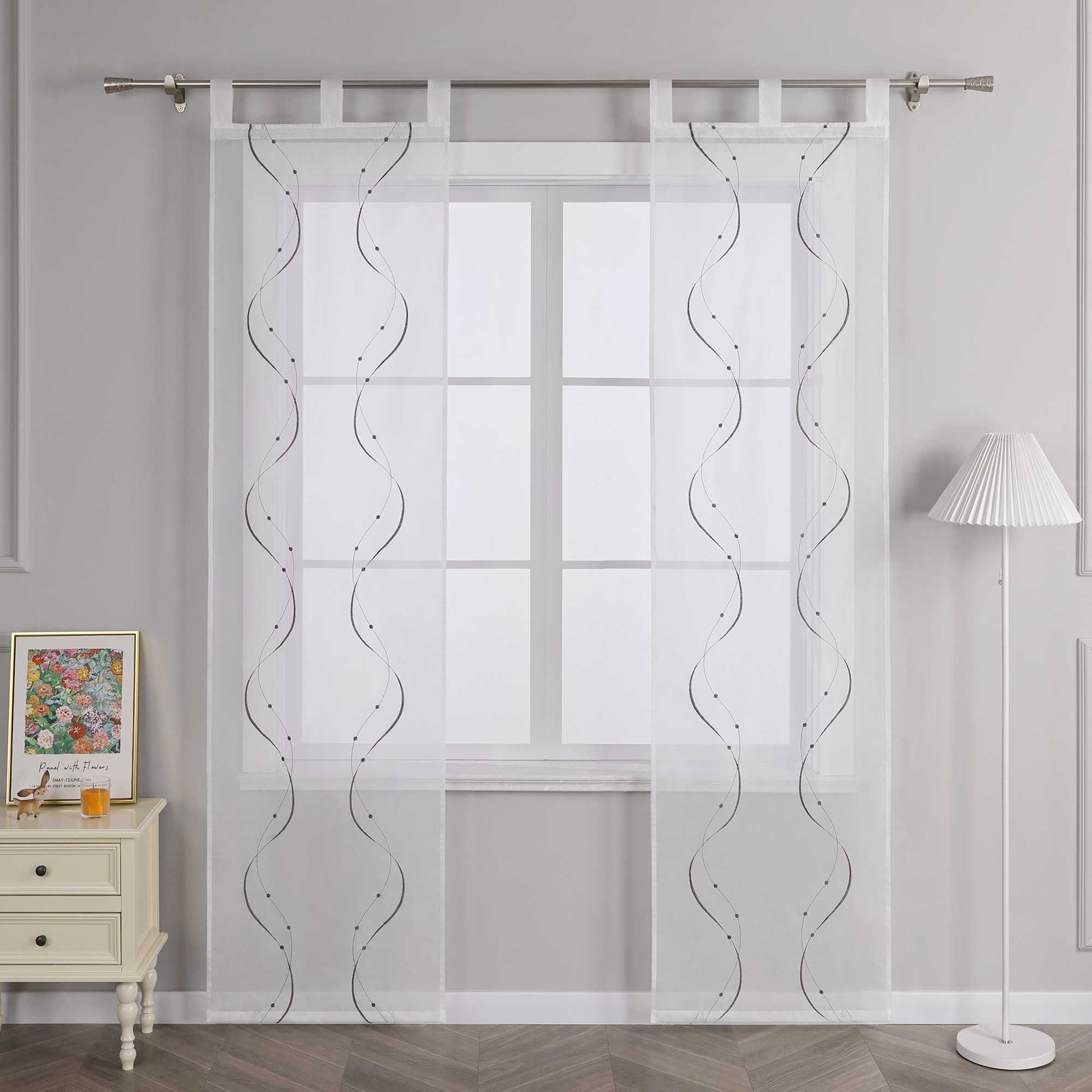 Vor einer hellgrauen Wand befindet sich ein Fenster, an dem eine weisse transparente Gardine mit grauem symmetrischem Wellenperlenmuster und Stickerei haengt. Die Gardine ist an einer Metallstange mit Schlaufen befestigt. Links vom Fenster steht ein heller Schrank mit Schubladen, darauf ein farbiges Blumenbild und ein kleines orangefarbenes Objekt. Rechts vom Fenster steht eine Stehlampe mit weissem gestreiftem Lampenschirm.