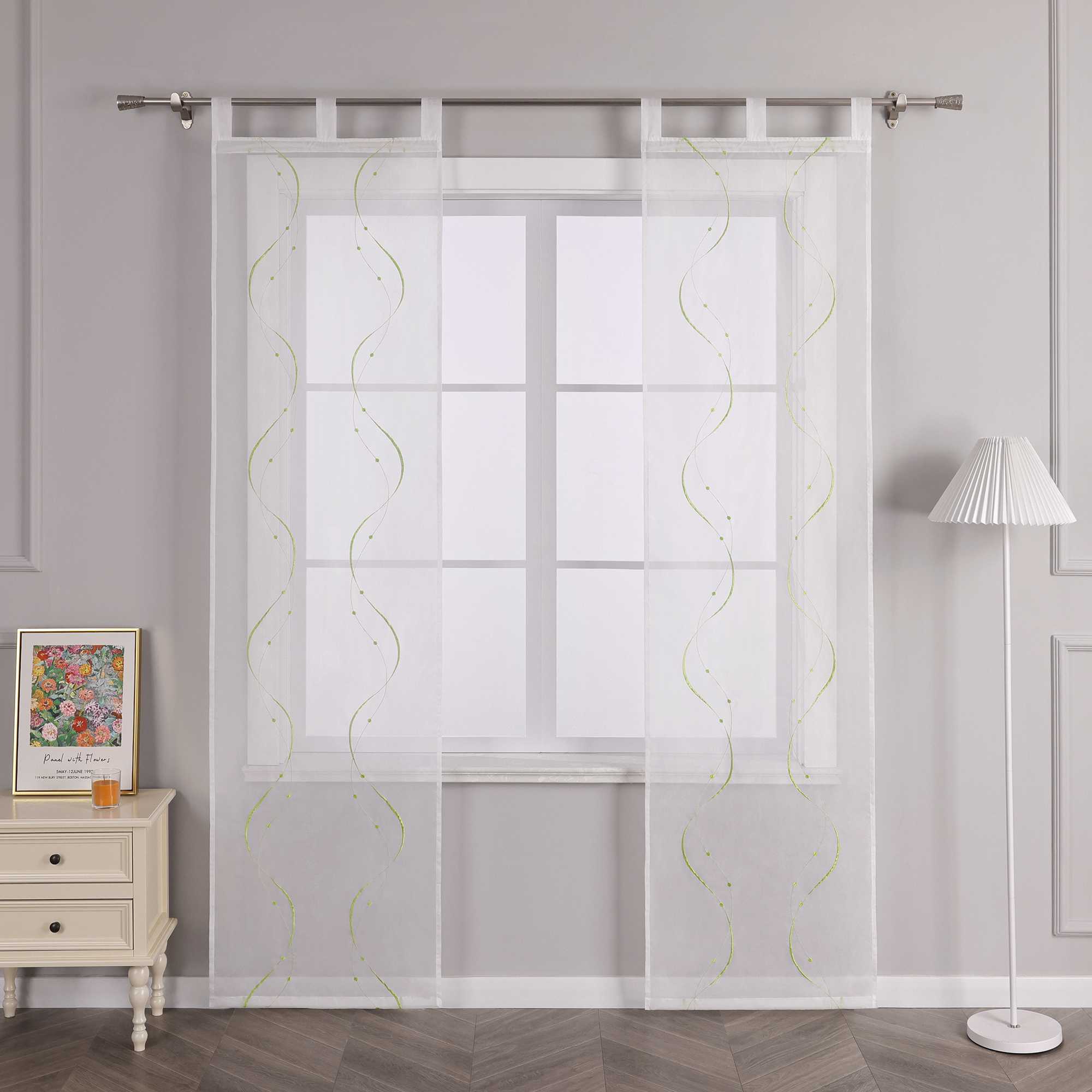 Vor einer hellgrauen Wand befindet sich ein Fenster, an dem eine weisse transparente Schiebegardine mit symmetrischem Wellenperlenmuster und grüner Stickerei haengt. Die Gardine ist an einer Metallstange mit Ringen befestigt. Links vom Fenster steht ein heller Schrank mit Schubladen, auf dem ein farbenfrohes Blumenbild und ein kleines oranges Objekt platziert sind. Rechts vom Fenster befindet sich eine Stehlampe mit weissem gestreiftem Lampenschirm.