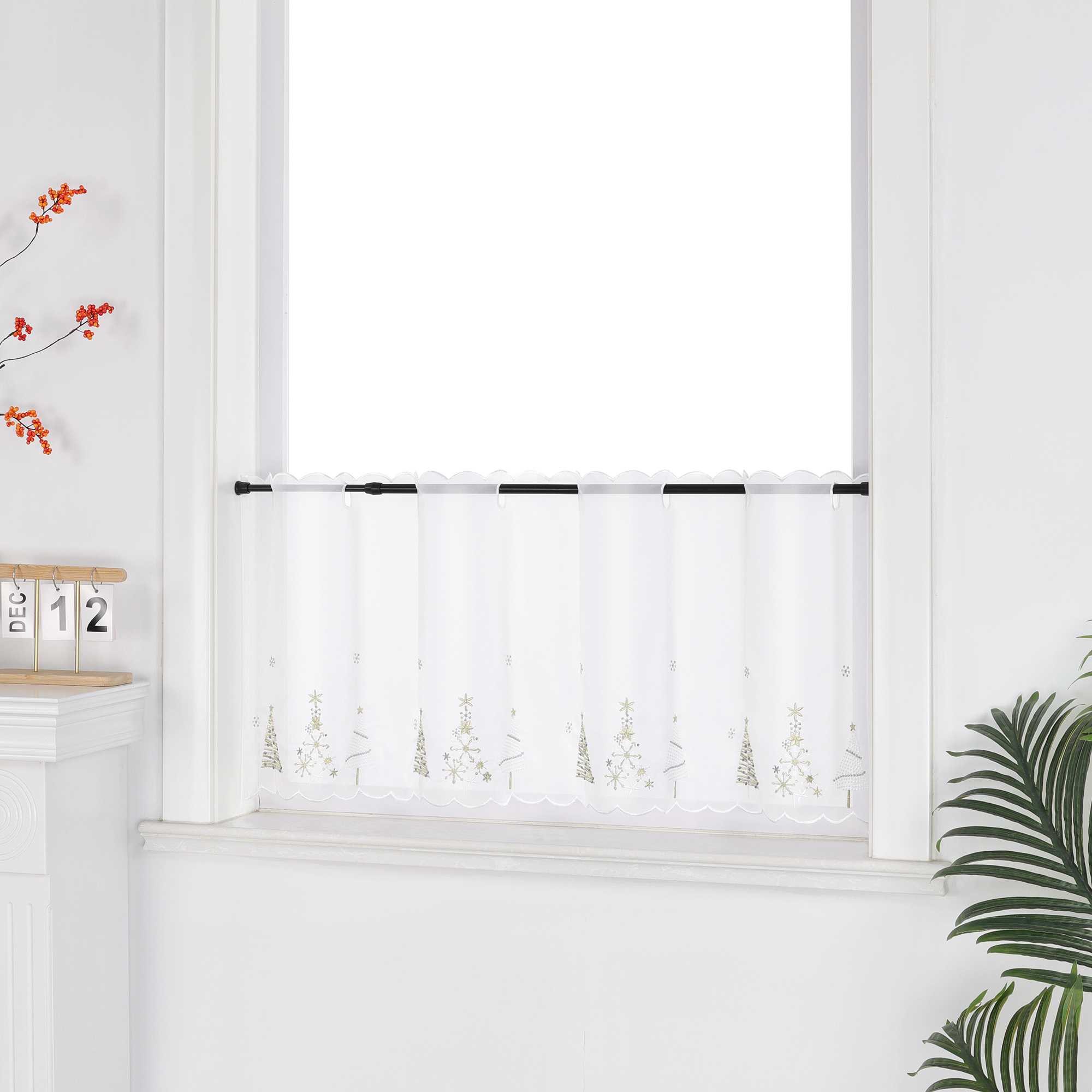 Eine weiße, transparente Scheibengardine mit goldenen Tannenbaum-Motiven hängt vor einem weißen Fenster. Die Wand ist ebenfalls weiß, was den hellen und festlichen Charakter unterstreicht. Seitlich sind dekorative Blumenarrangements zu sehen, die dem Bild eine natürliche und stimmungsvolle Note verleihen – ideal als stimmige Weihnachtsdekoration.