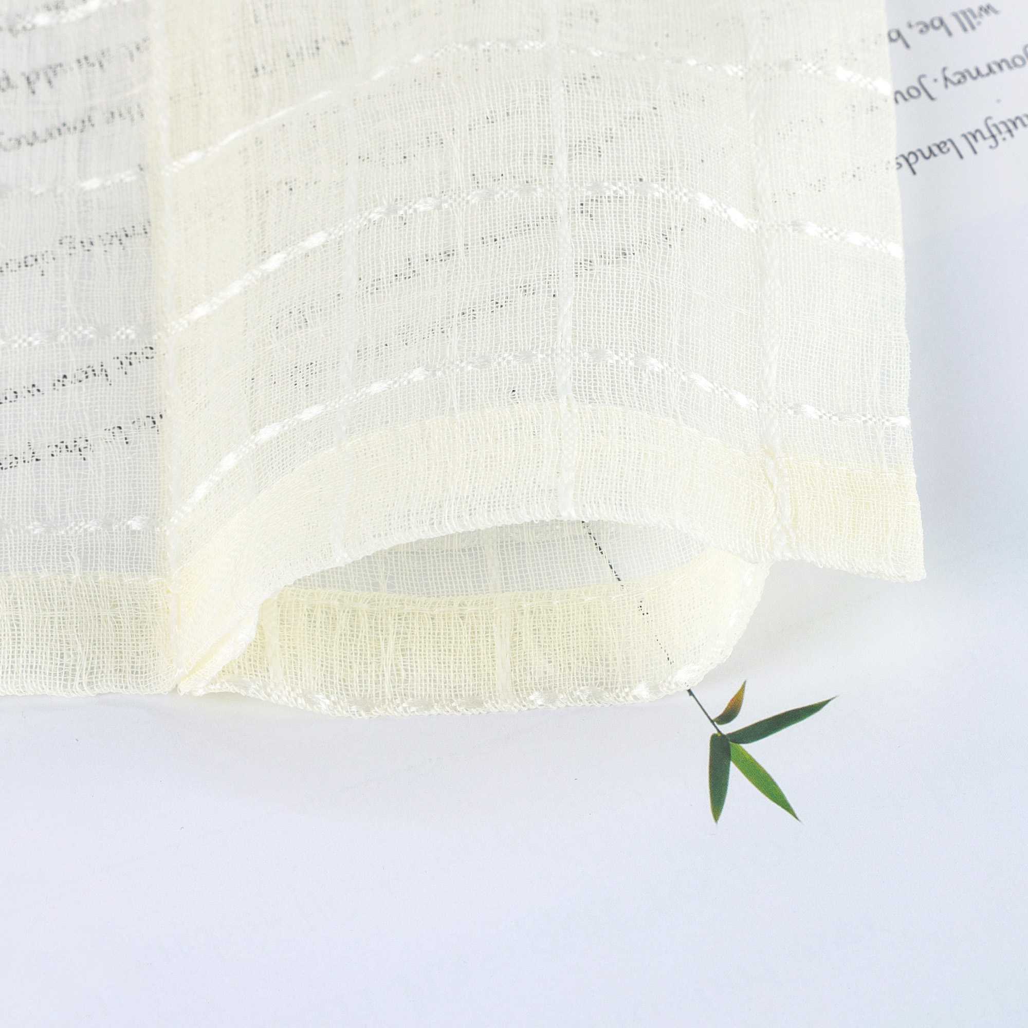 Der cremeweiße Vorhangstoff liegt locker gebogen auf einer hellen Fläche. Die feine Karostruktur und luftige Transparenz sind deutlich erkennbar. Ein grünes Blatt unter dem Stoff betont die weiche Textur und Lichtdurchlässigkeit.