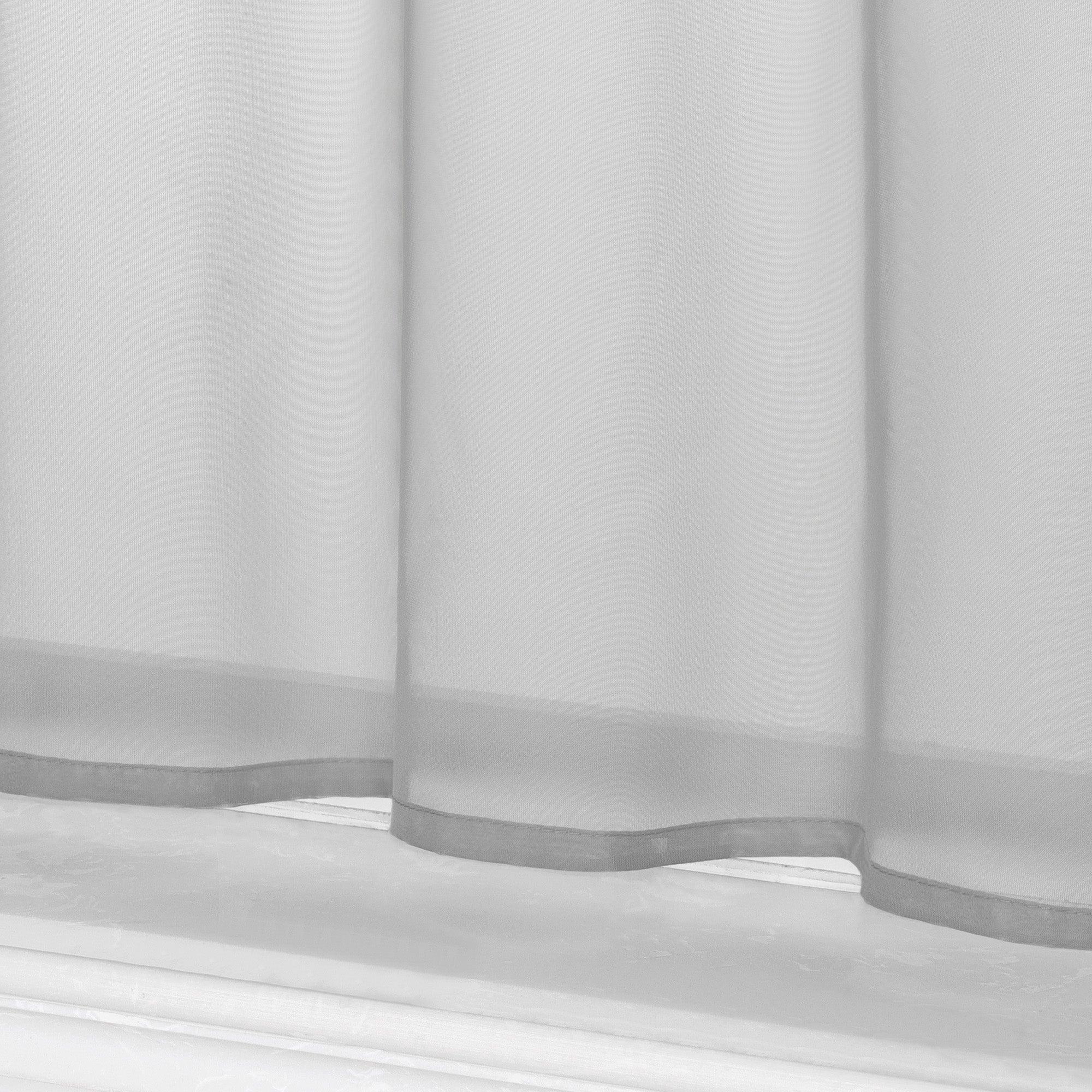 Dies ist eine Detailaufnahme des unteren Teils des grauen kurzen Vorhangs. Der Vorhang zeigt das leichte, halbtransparente Material sowie den wellenförmigen Saum am unteren Rand. Darunter ist die Fensterbank zu sehen.