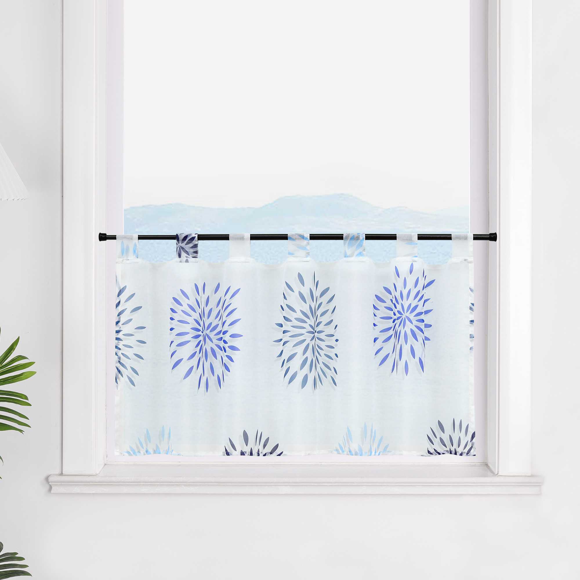 Die transparente Scheibengardine mit blauen Feuerwerksmotiven hängt vor einem Fenster. Der weiße Hintergrund lässt die blauen Muster des Feuerwerks strahlen und sorgt für eine festliche, aber beruhigende Atmosphäre im Raum.