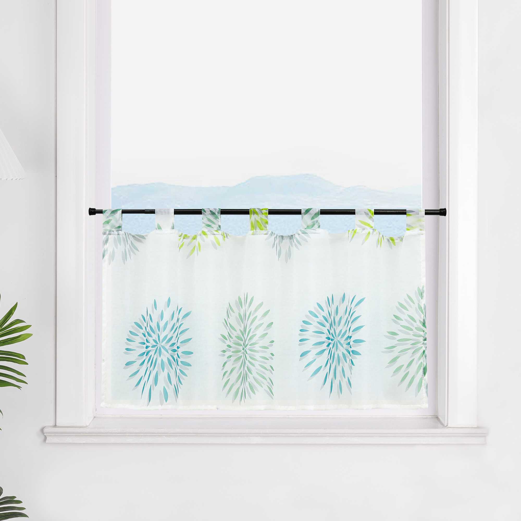 Die transparente Scheibengardine mit grünen und blauen Feuerwerksmotiven hängt dekorativ vor einem Fenster. Die Wand im Hintergrund ist in weiß gehalten, wodurch die bunten Feuerwerksmuster auf dem Stoff besonders zur Geltung kommen. Die Gardine verleiht dem Raum eine festliche, fröhliche Atmosphäre.