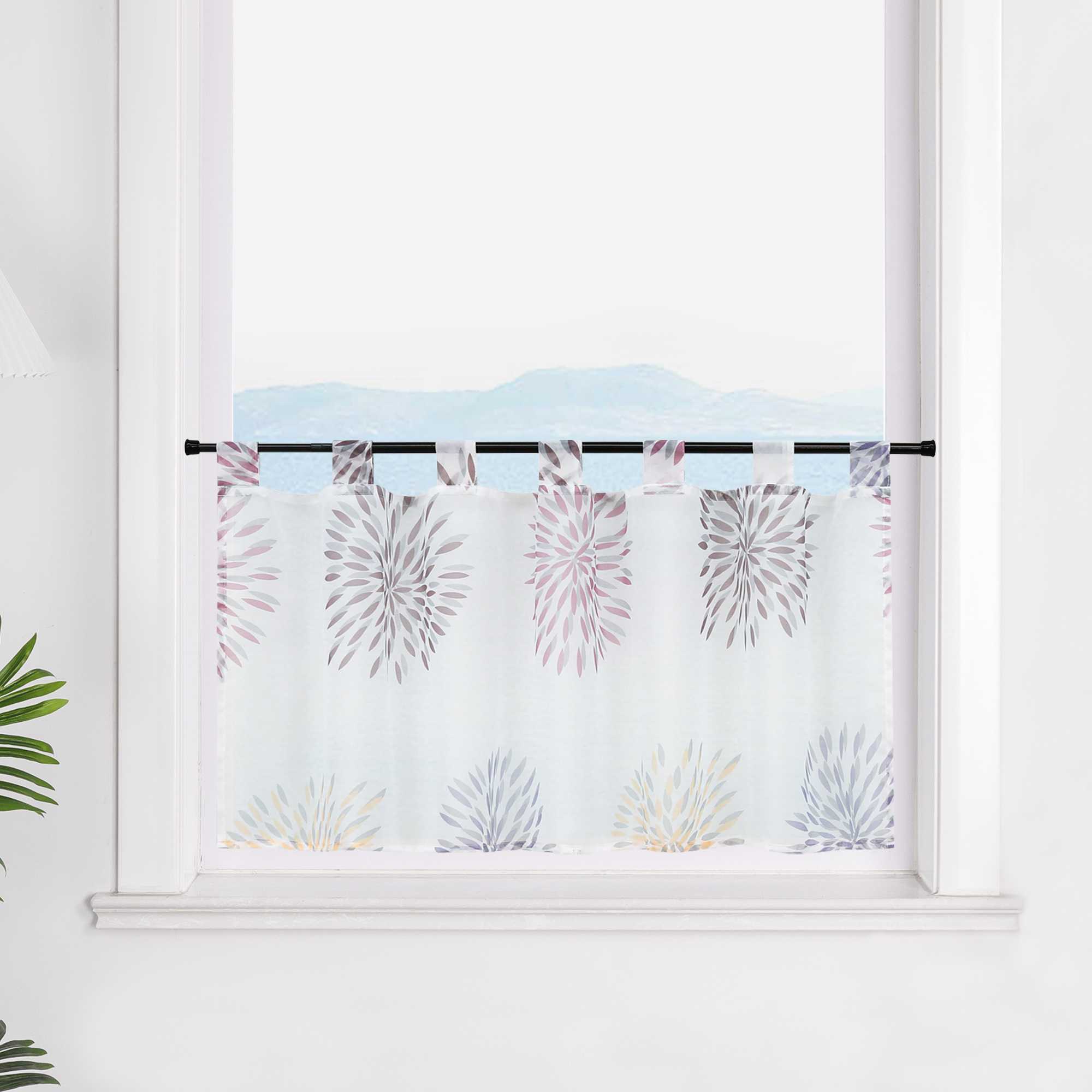 Die transparente Scheibengardine mit lila Feuerwerksmotiven hängt vor einem Fenster. Die weiße Wand im Hintergrund hebt die lila Muster des Feuerwerks hervor und verleiht dem Raum eine festliche und elegante Stimmung.