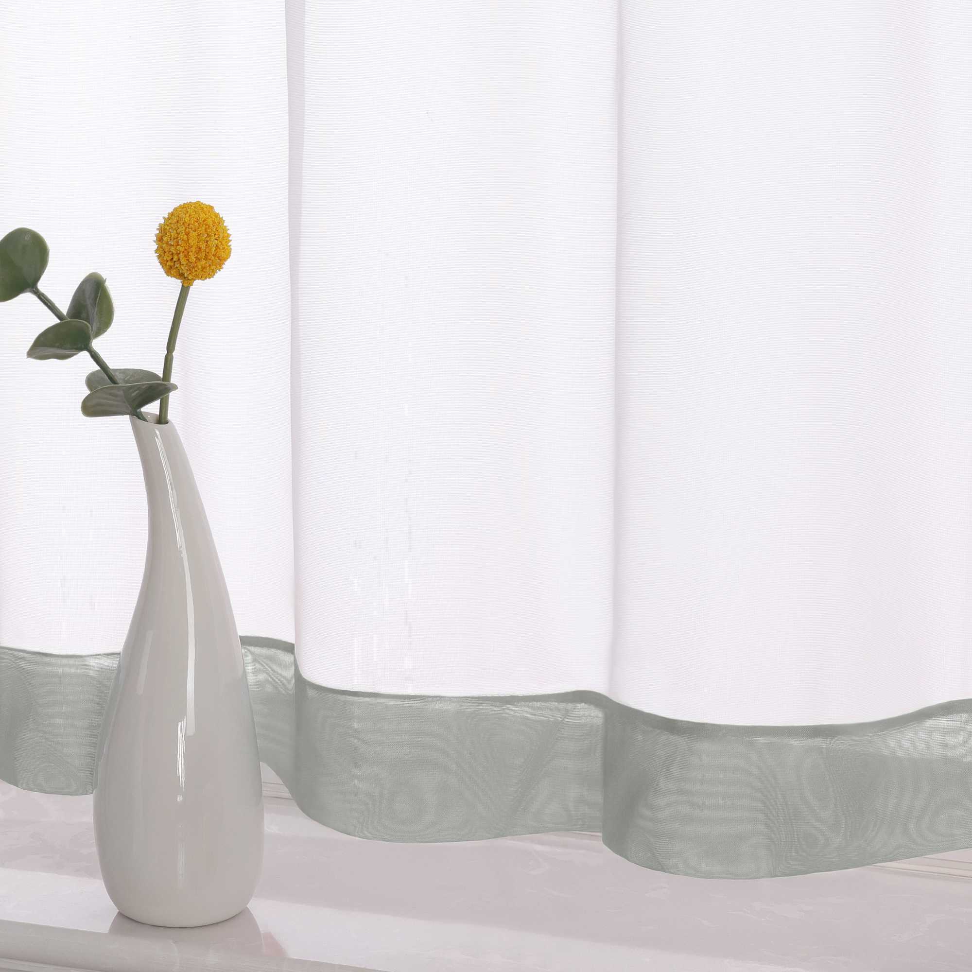 Die Unterkante des Vorhangs wird im Detail gezeigt, mit einem grauen Dekorband als stilvollem Akzent. Ein Vase im Vordergrund ergänzt die Szene und fügt dem Raum eine harmonische, dekorative Note hinzu, die den eleganten Look des Vorhangs unterstreicht.