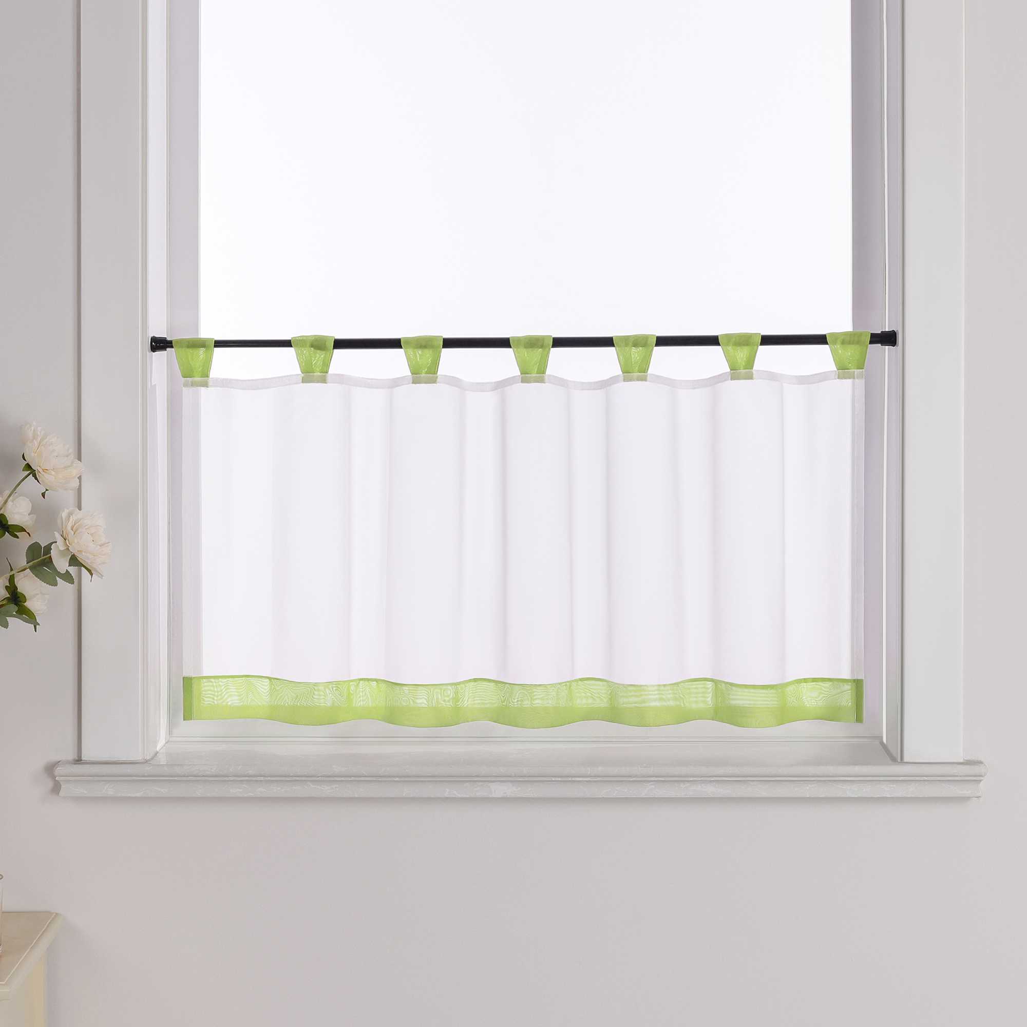 Die gruene Scheibengardine hängt vor einem Fenster, wobei die Hintergrundwand weiß ist. Der Vorhang ist transparent und wird von blumigen Dekorationen an der Seite begleitet.
