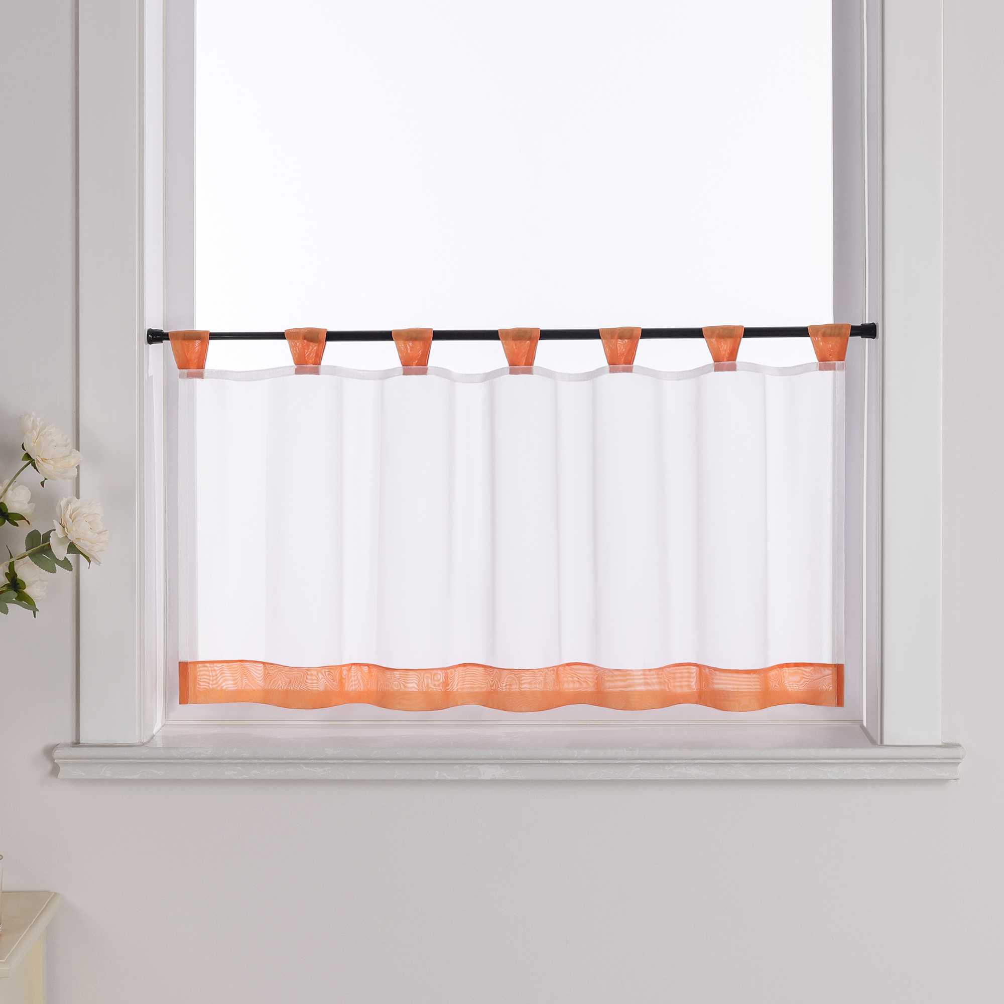 Die orange Scheibengardine hängt vor einem Fenster, wobei die Hintergrundwand weiß ist. Der Vorhang ist transparent und wird von blumigen Dekorationen an der Seite begleitet.