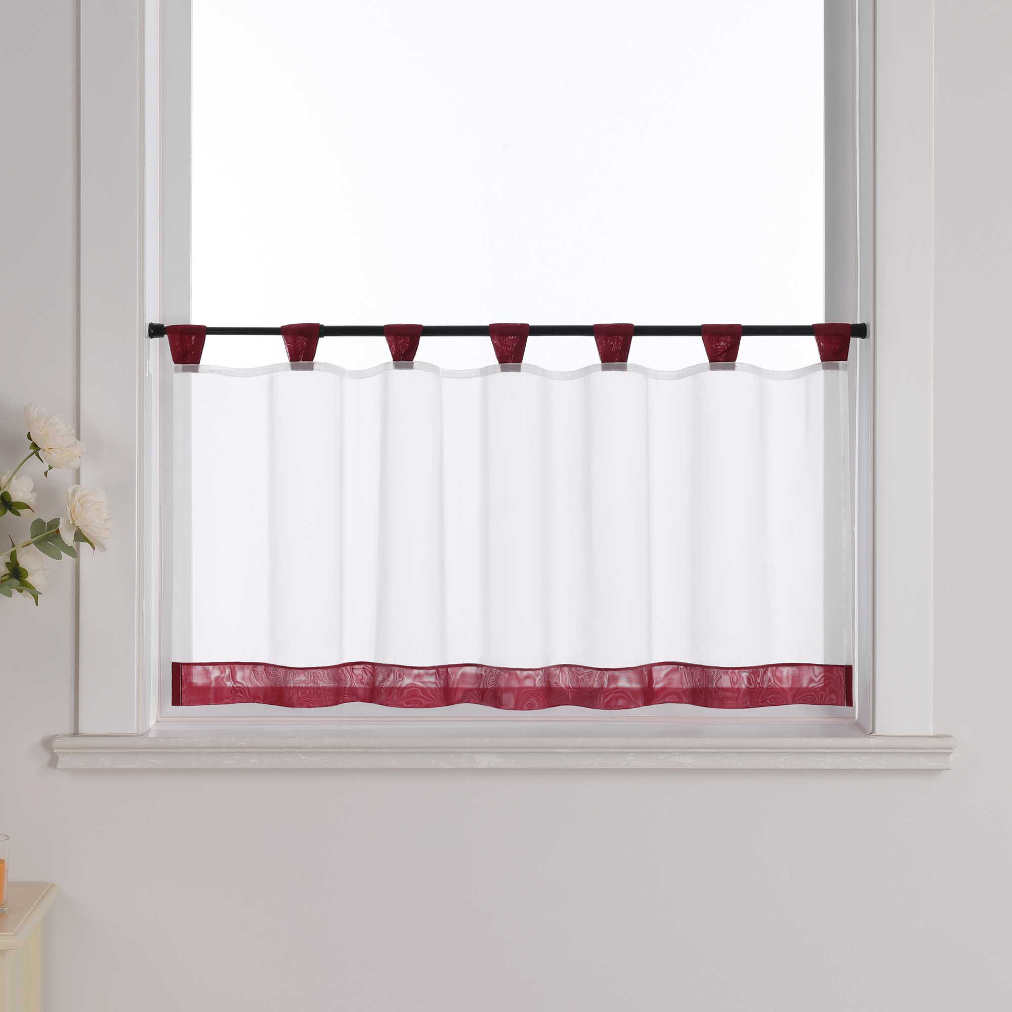 Die weinrot Scheibengardine hängt vor einem Fenster, wobei die Hintergrundwand weiß ist. Der Vorhang ist transparent und wird von blumigen Dekorationen an der Seite begleitet.